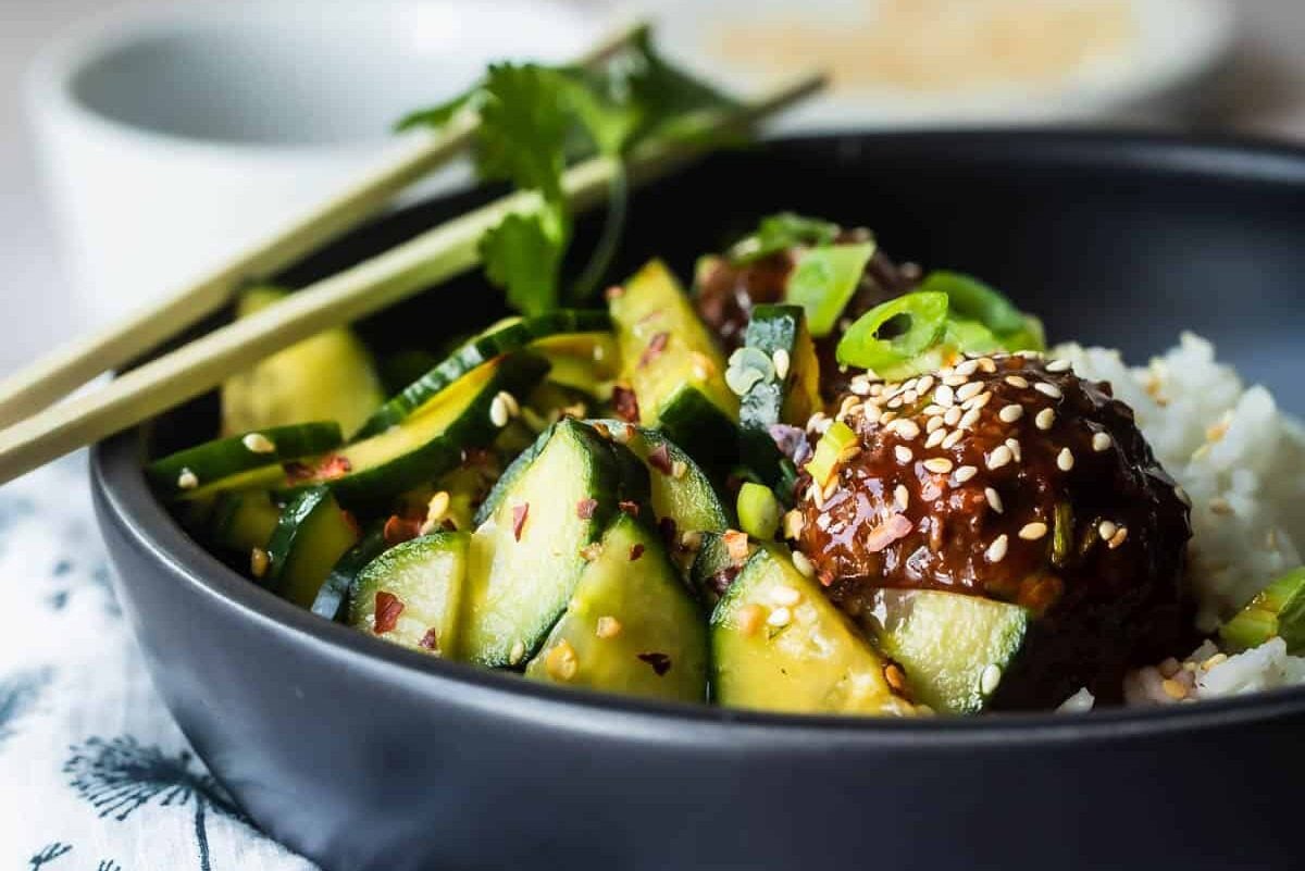 A black bowl of Asian cucumber salad, meatballs and rice.