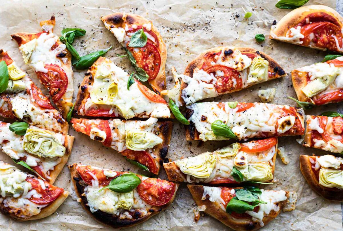 Flatbread pizzas topped with artichokes, tomatoes, cream cheese, and basil leaves on a board.