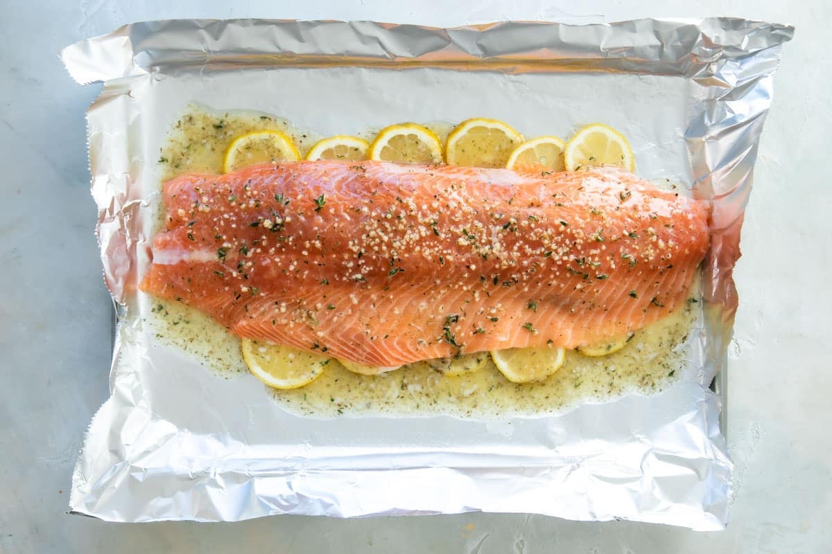 A large side of raw salmon with spices on top sitting on a layer of lemon slices on top of foil on a baking sheet.
