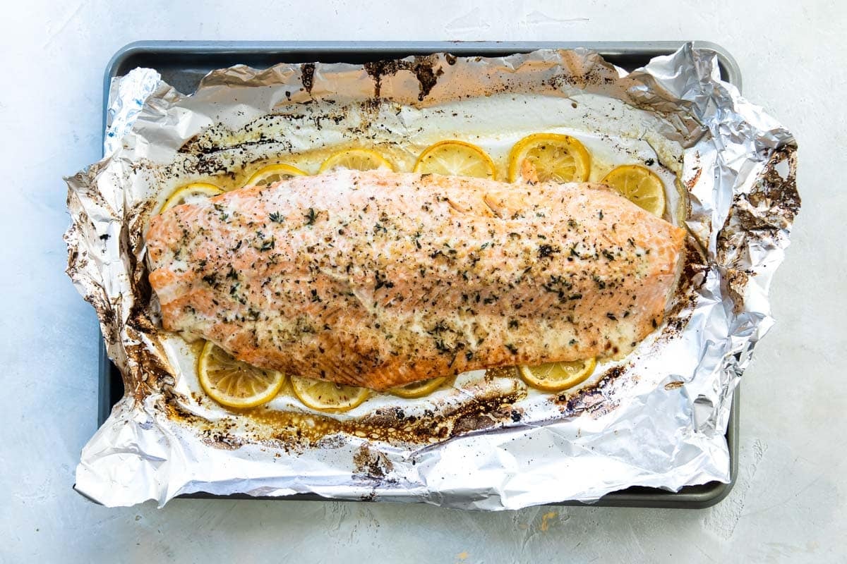 A large side of cooked salmon with spices on top sitting on a layer of lemon slices on top of foil on a baking sheet.