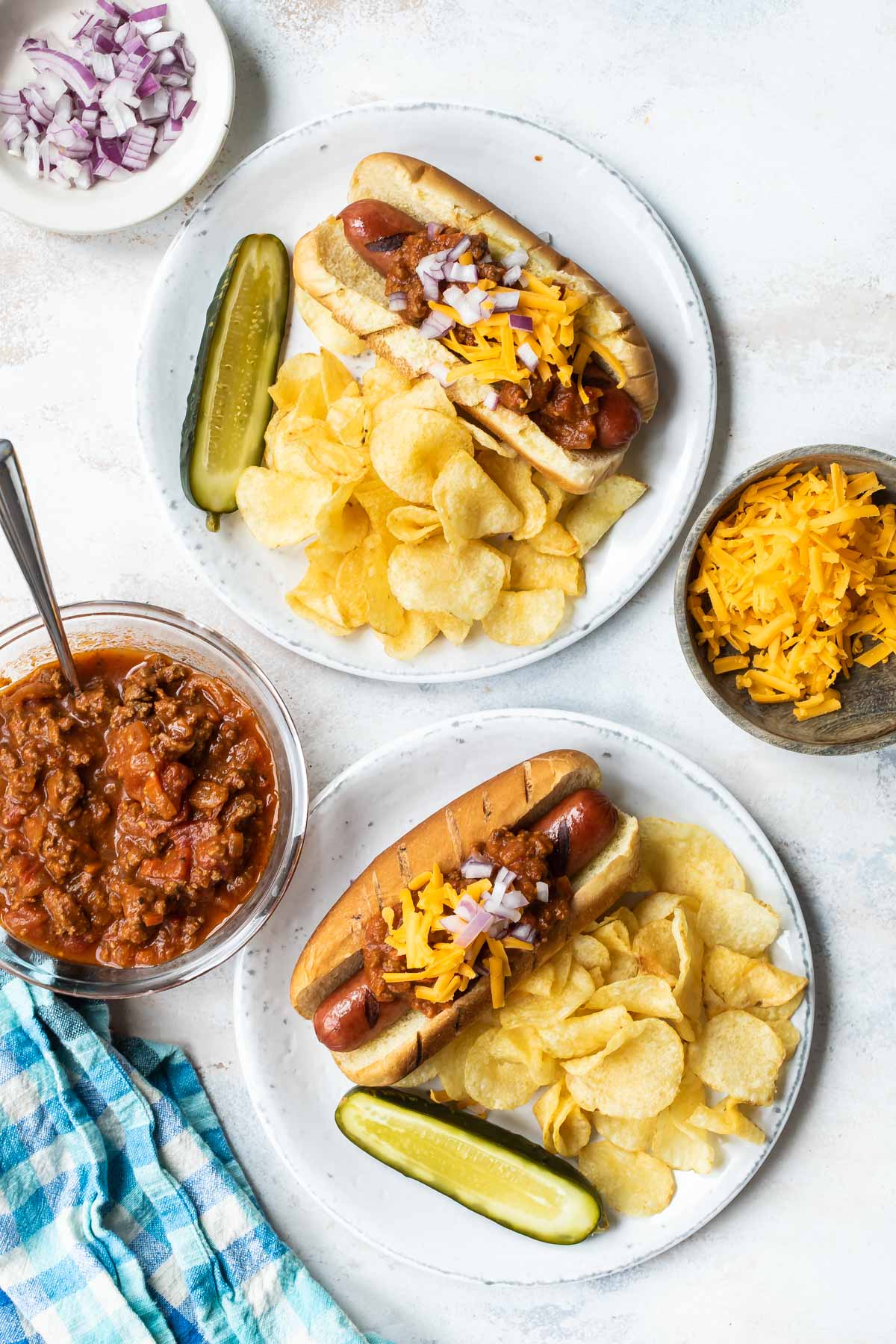 Two white plates with chili dogs and potato chips next to a bowl of Hot Dog Chili.