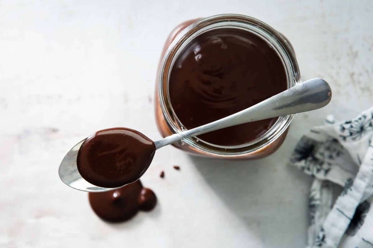 Chocolate sauce on a spoon that is resting over a mason jar of chocolate sauce.