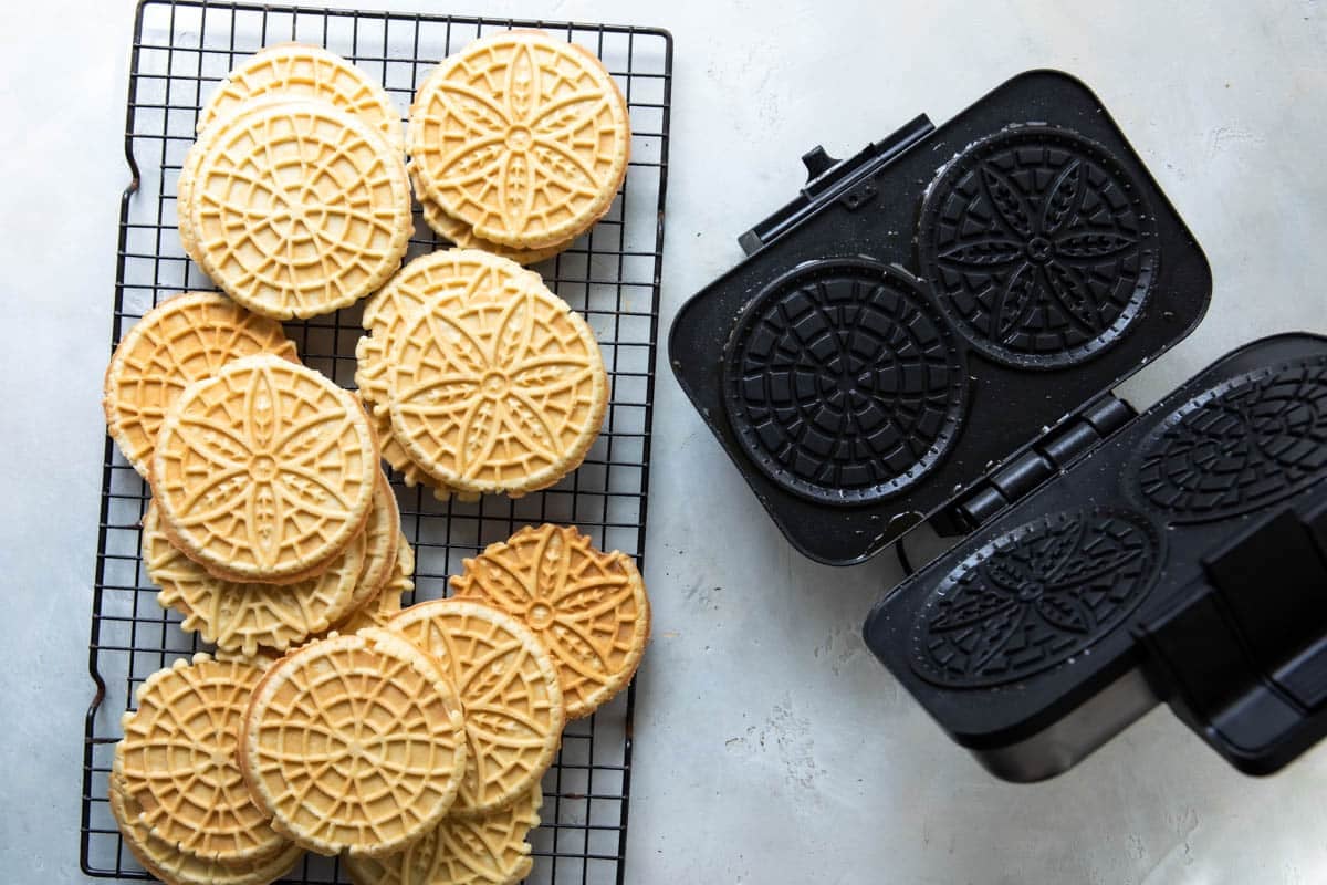 Pizzelle cookies cooling on a baking rack next to a pizzelle iron.