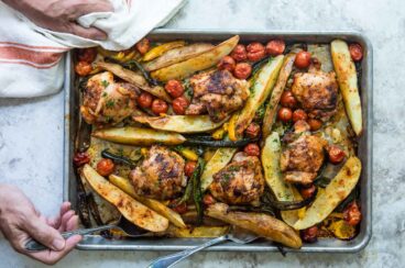 Someone scooping a potato off of a sheet pan filled with vegetables and paprika chicken.