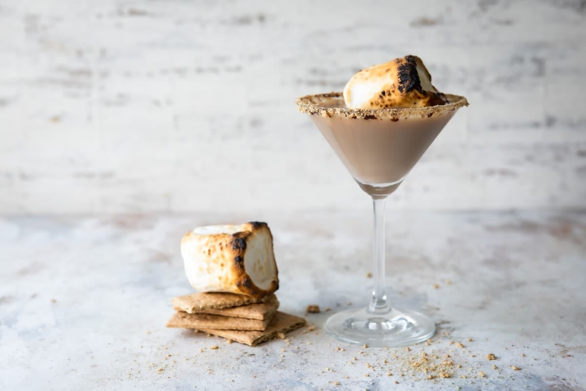 A S'mores Martini in a martini glass next to some graham crackers and a toasted marshmallow.