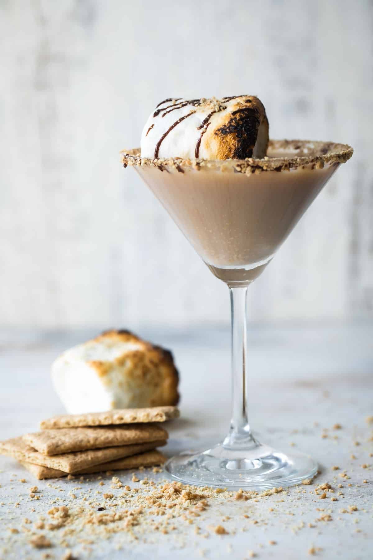 A S'mores Martini in a martini glass next to some graham crackers and a toasted marshmallow.