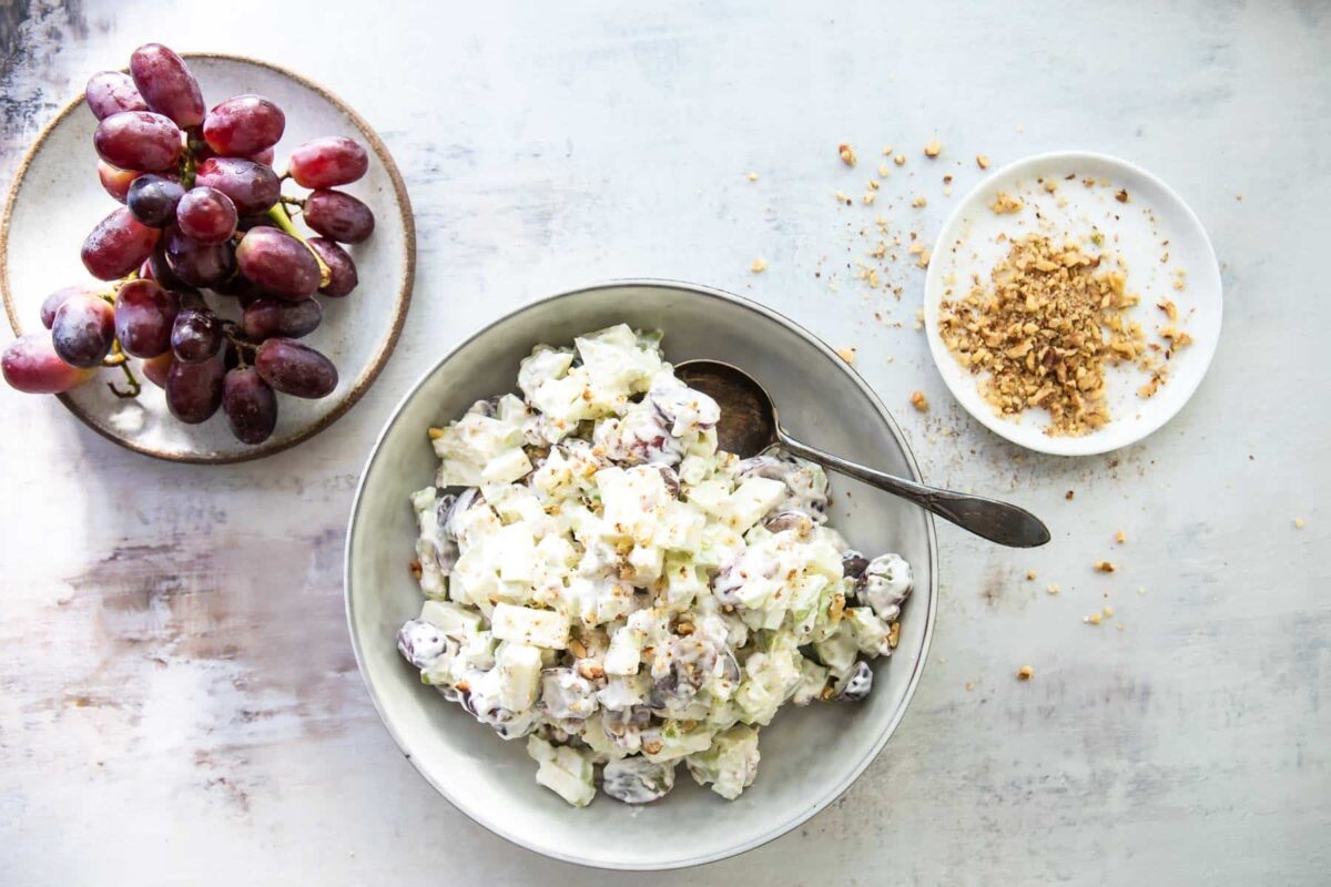 A white serving bowl with waldorf salad in it next to a plate of grapes.