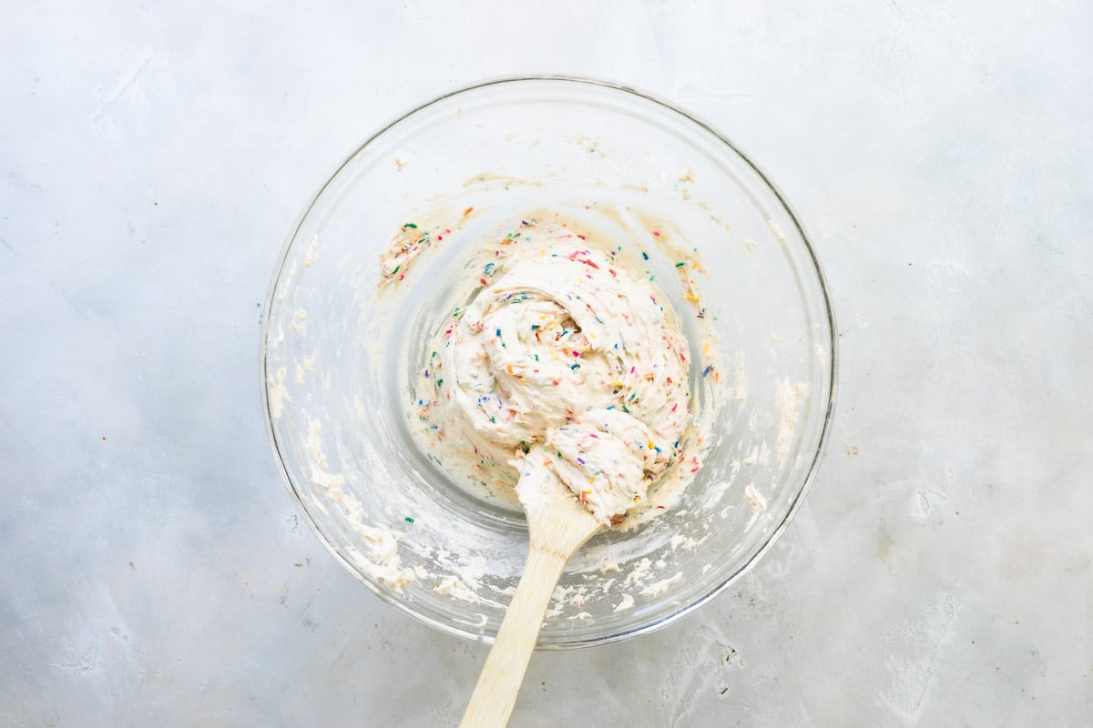 Confetti cookie dough in a clear bowl with a wooden spoon.