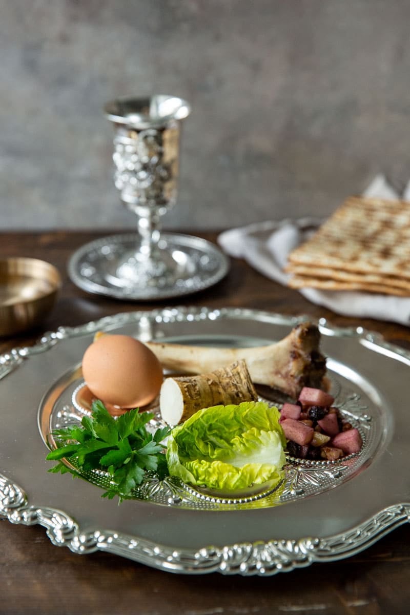 A silver Seder Plate with the seder components and a cup of wine and Matzah in the background.