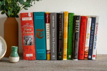 A collection of 12 cookbooks lined up together on a cabinet below a mirror.