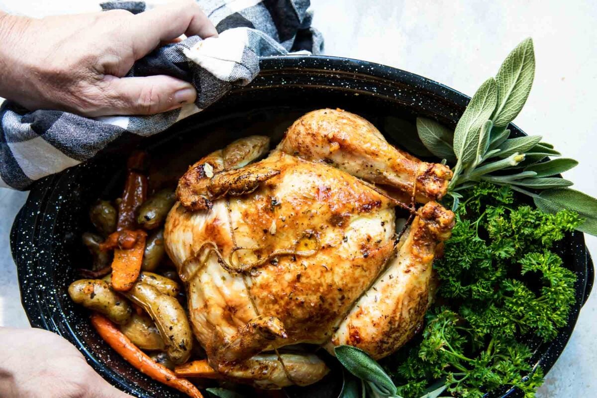 Someone holding a garlic butter roast chicken in a black roasting pan.