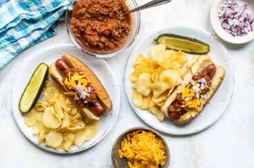 Two plates with chili dogs, chips and a pickle.