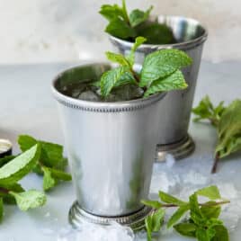Two mint juleps in silver cups with a bottle of whiskey in the background.