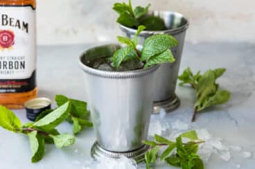 Two mint juleps in silver cups with a bottle of whiskey in the background.