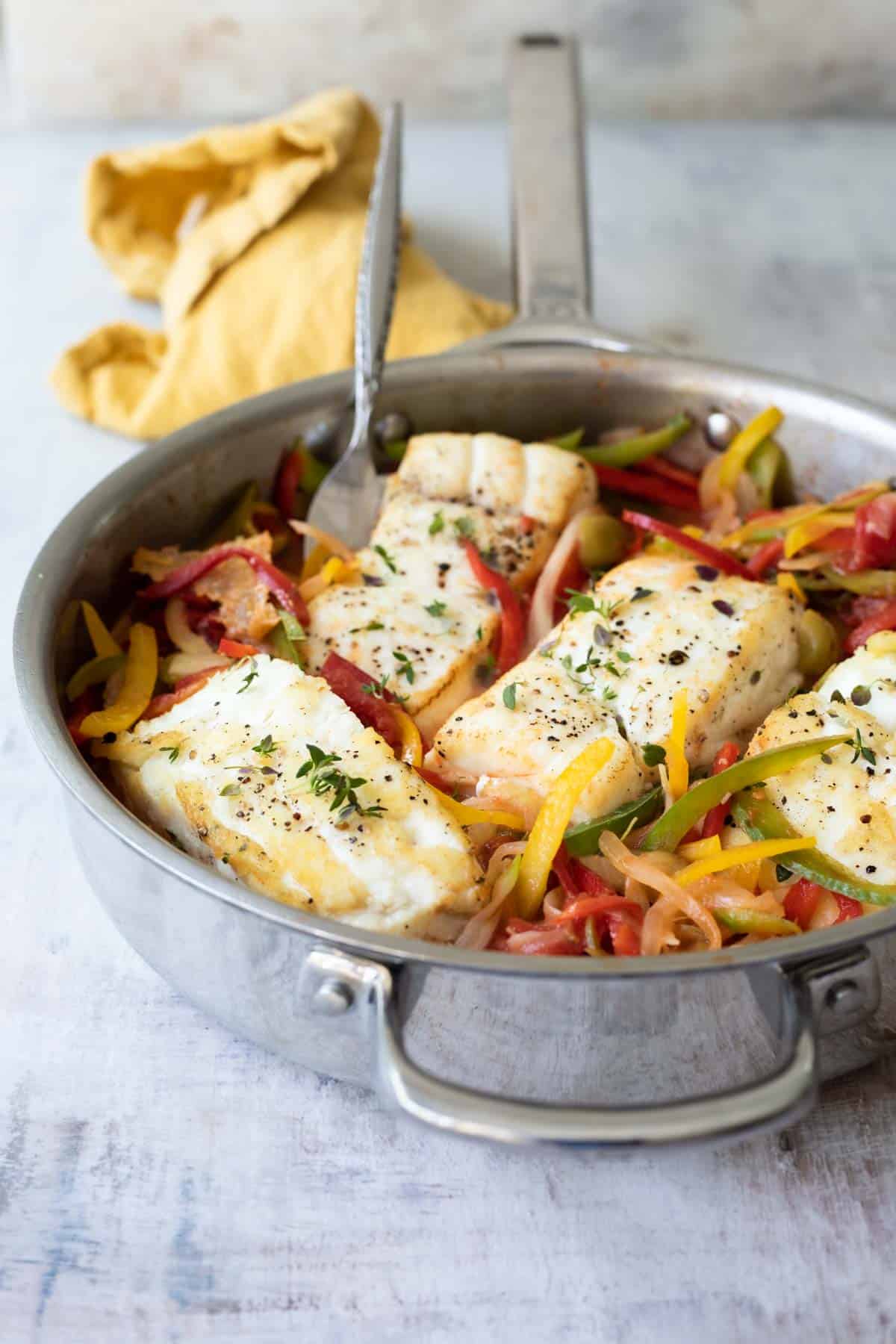 A skillet full of seared halibut, sliced bell peppers and green olives.