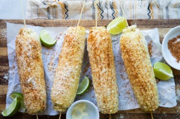 Four cobs of Mexican Street Corn on a paper on a cutting board surrounded by limes.