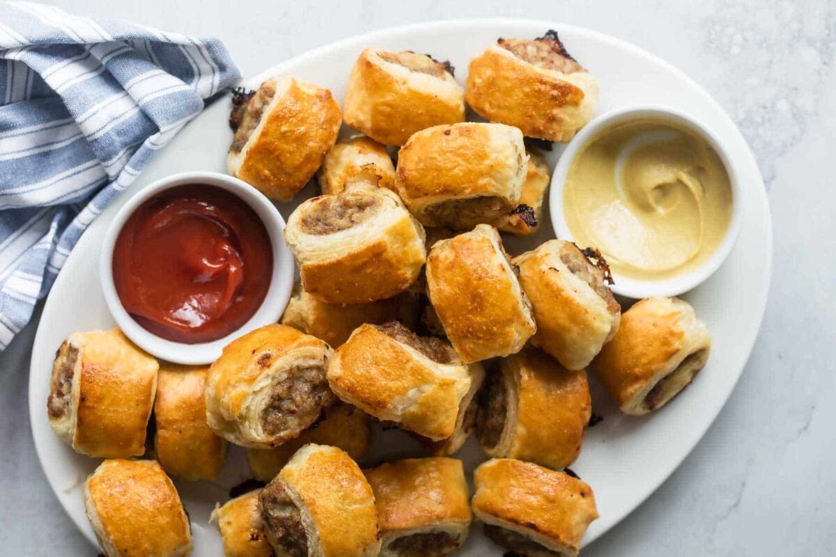 A platter with sausage rolls and two condiment cups containing ketchup and mustard.