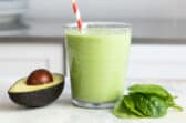 A glass of avocado smoothie next to a pile of spinach and half of an avocado.