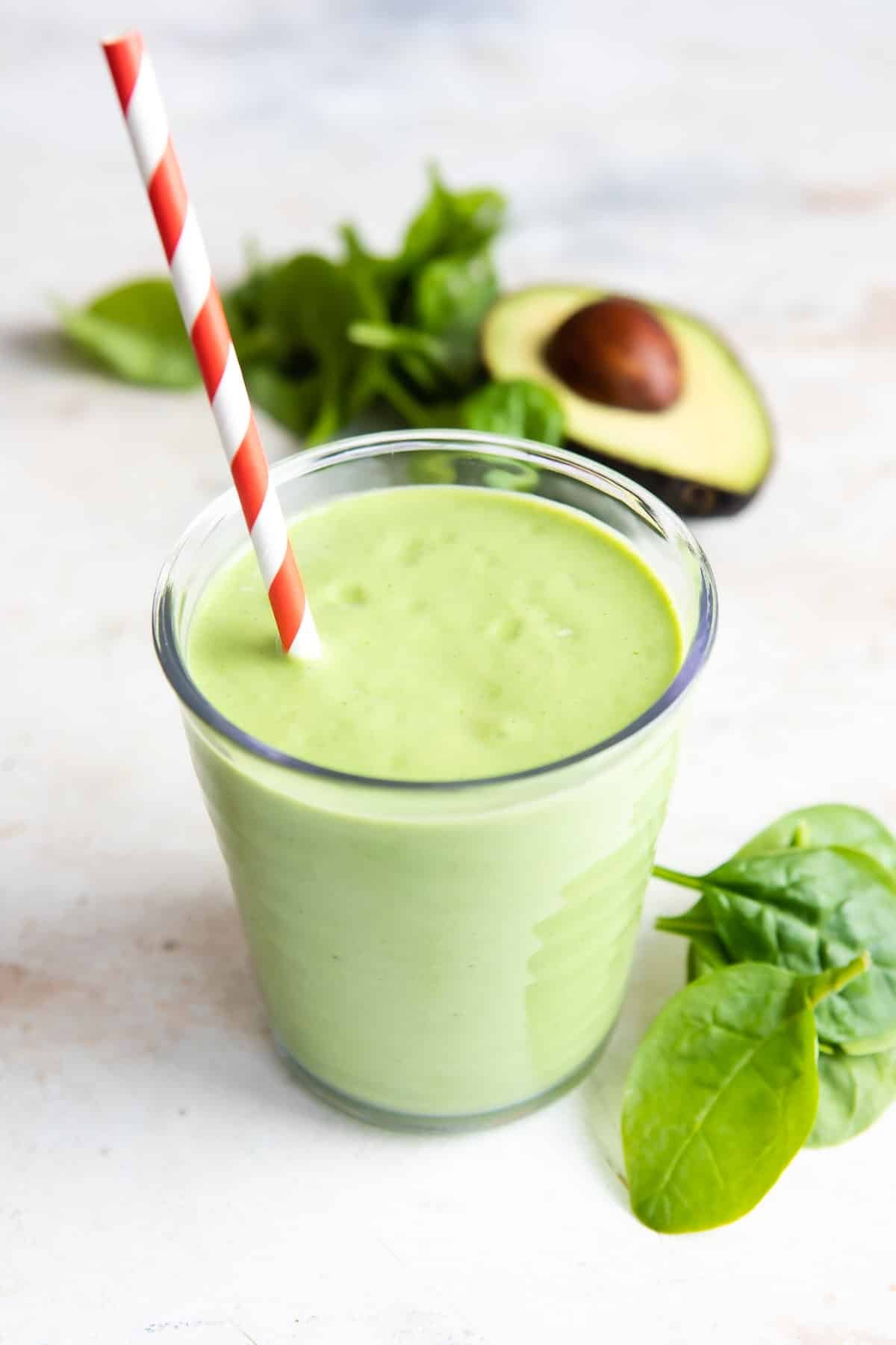 A glass of avocado smoothie next to a pile of spinach and half of an avocado.