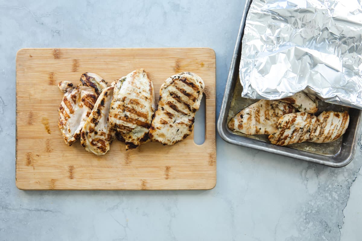 Some grilled lemon chicken on a pan tented with foil, other pieces on a cutting board.
