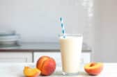 A glass of peach smoothie next to fresh peaches on a counter.