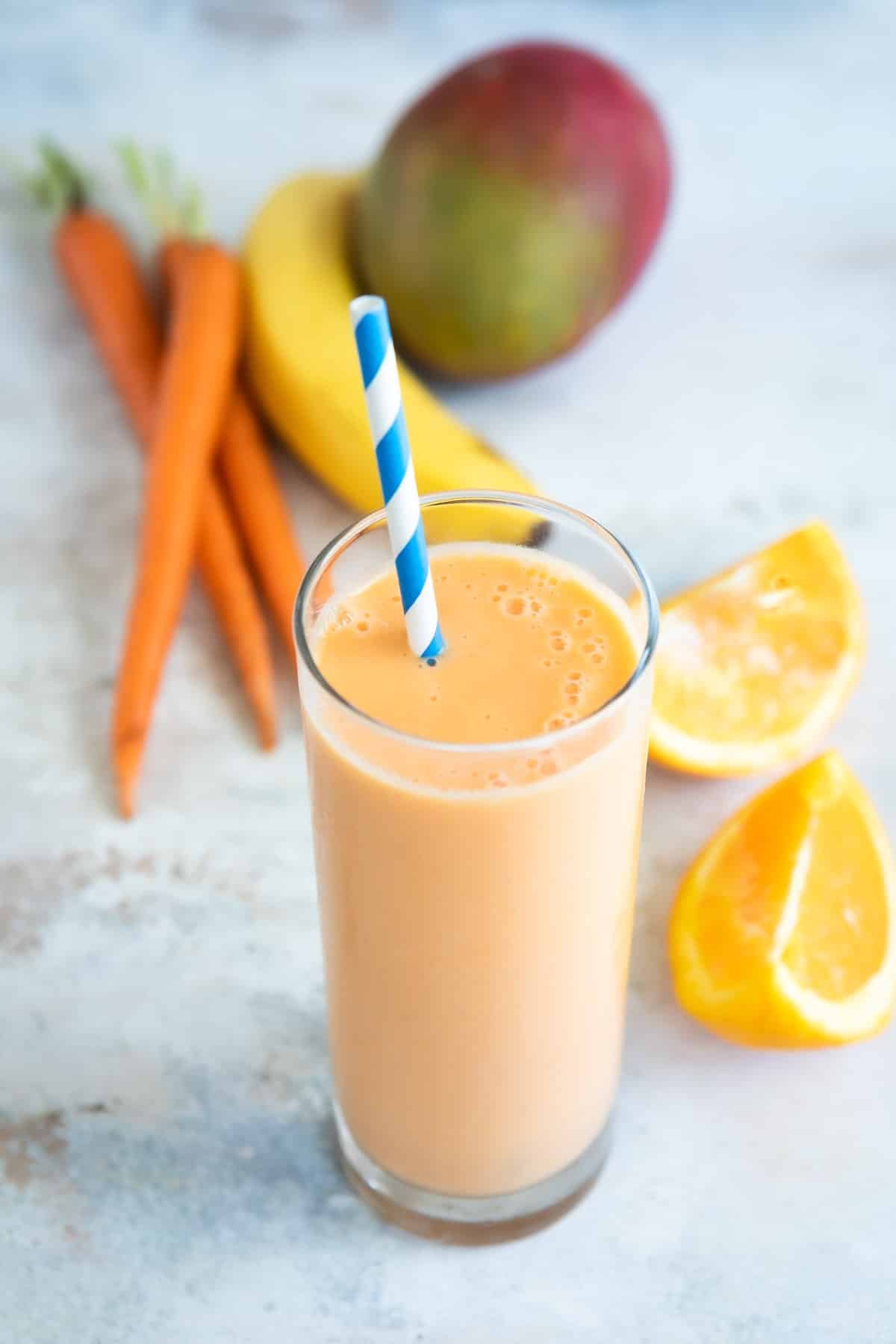 A glass of carrot smoothie next to a mango, carrots, and a banana.