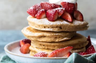 A stack of healthy pancakes with strawberries, whipped cream and powdered sugar.