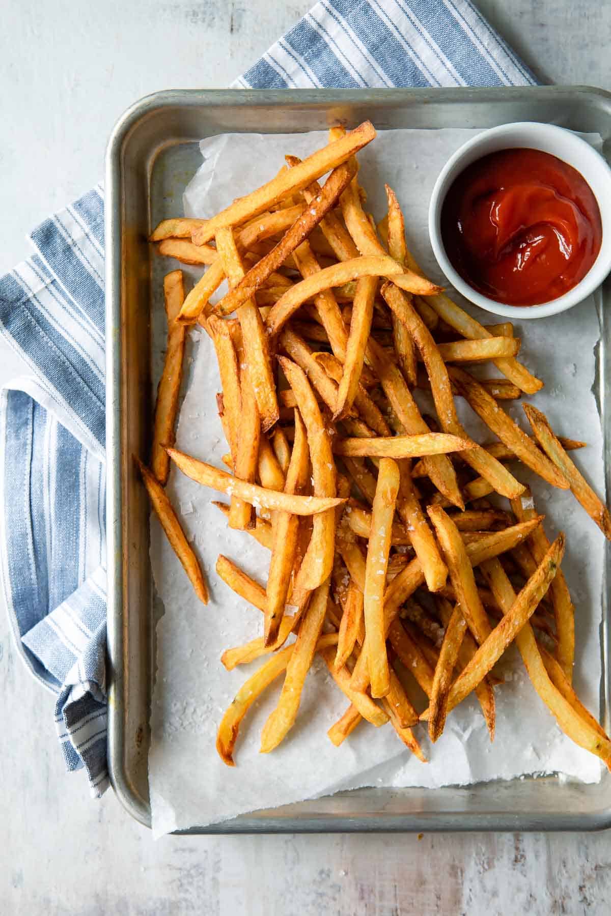 A pile of french fries on a baking sheet next to a cup of ketchup.