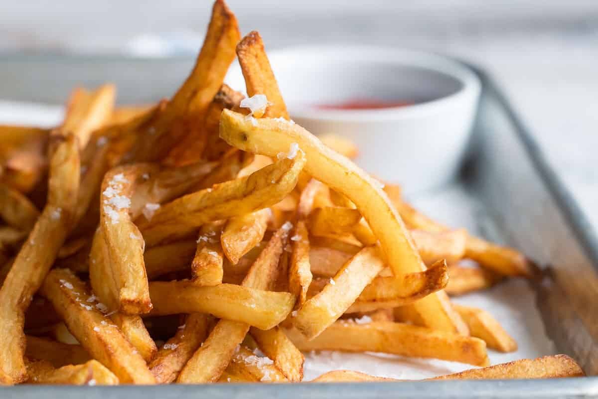 A pile of french fries on a baking sheet next to a cup of ketchup.