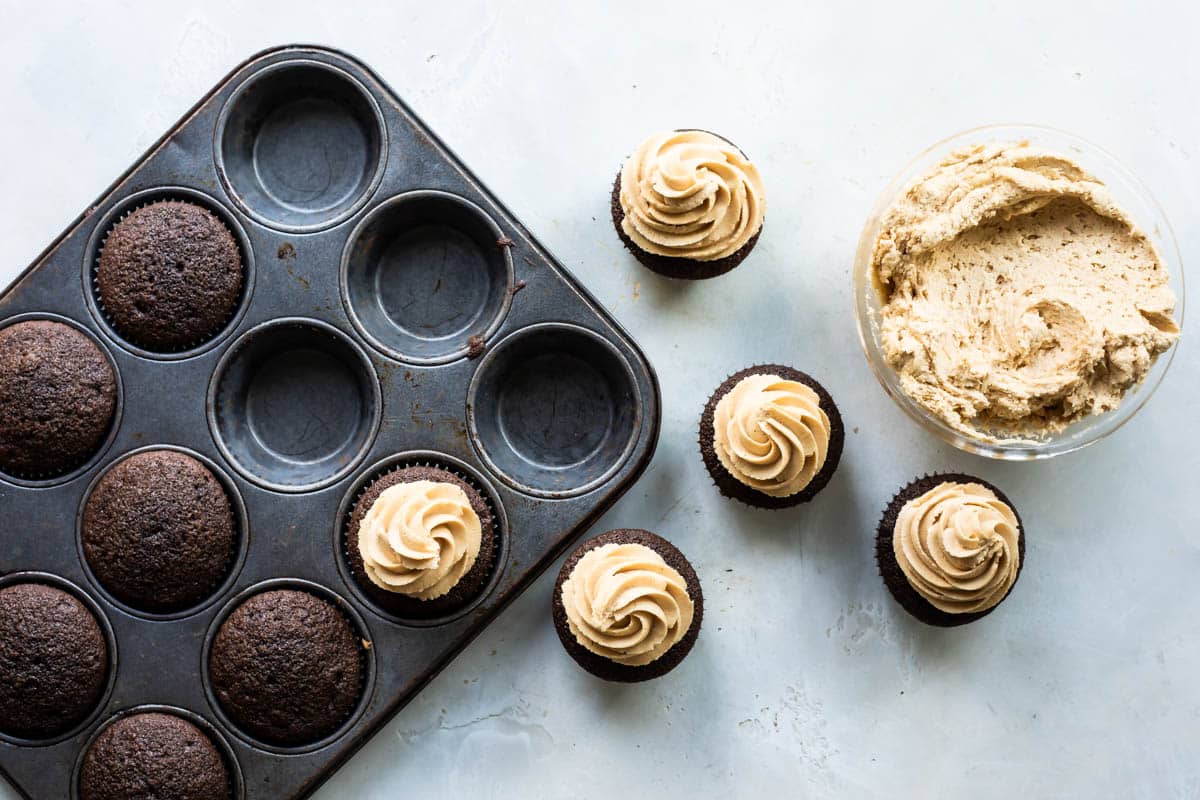 A muffin tin with chocolate cupakes inside next to a bowl of peanut butter frosting.