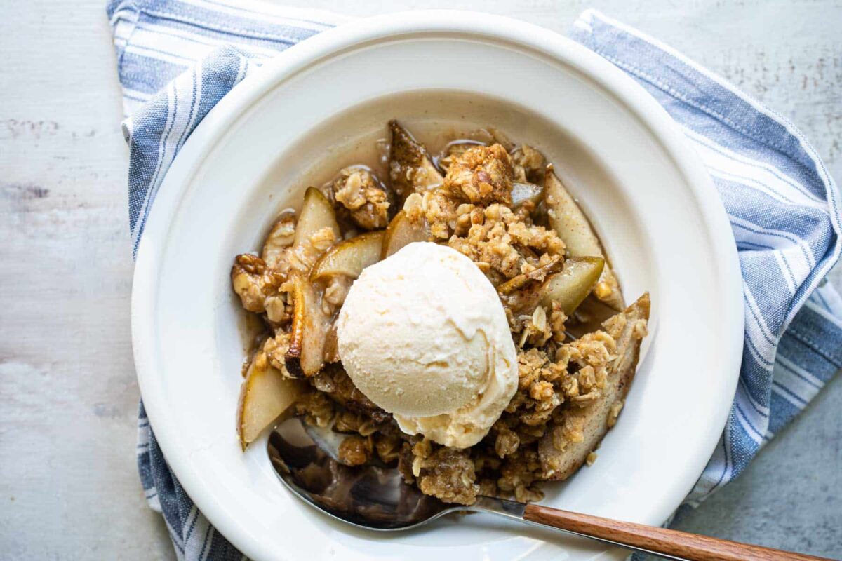 A white bowl of pear crisp with vanilla ice cream on top.