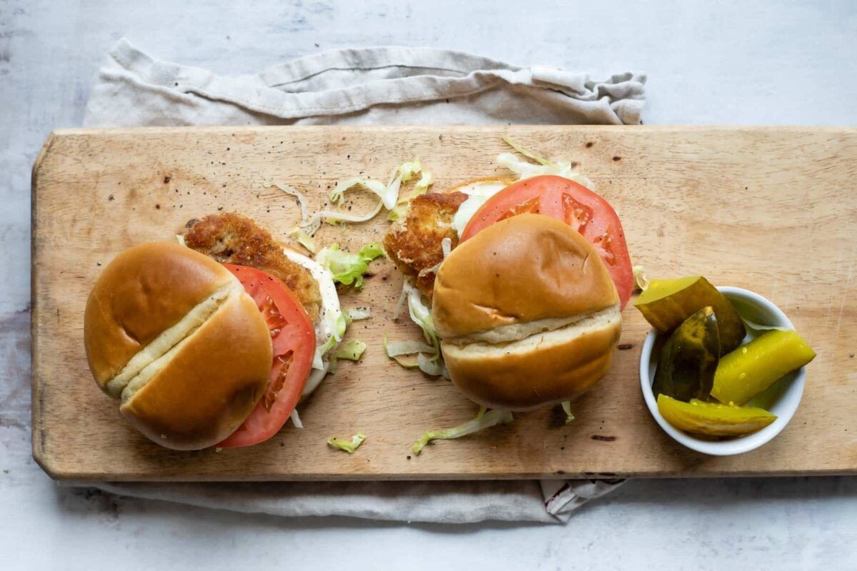 2 pork tenderloin sandwiches on a board next to a dish of pickles.
