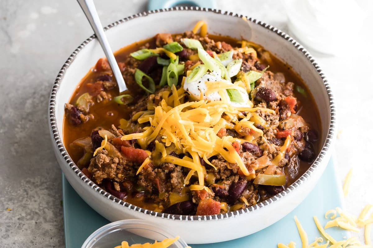 Beef chili in a white bowl on a teal cutting board surrounded by shredded cheese.