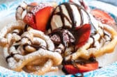 A funnel cake topped with strawberries, ice cream, and chocolate syrup on a paper plate.