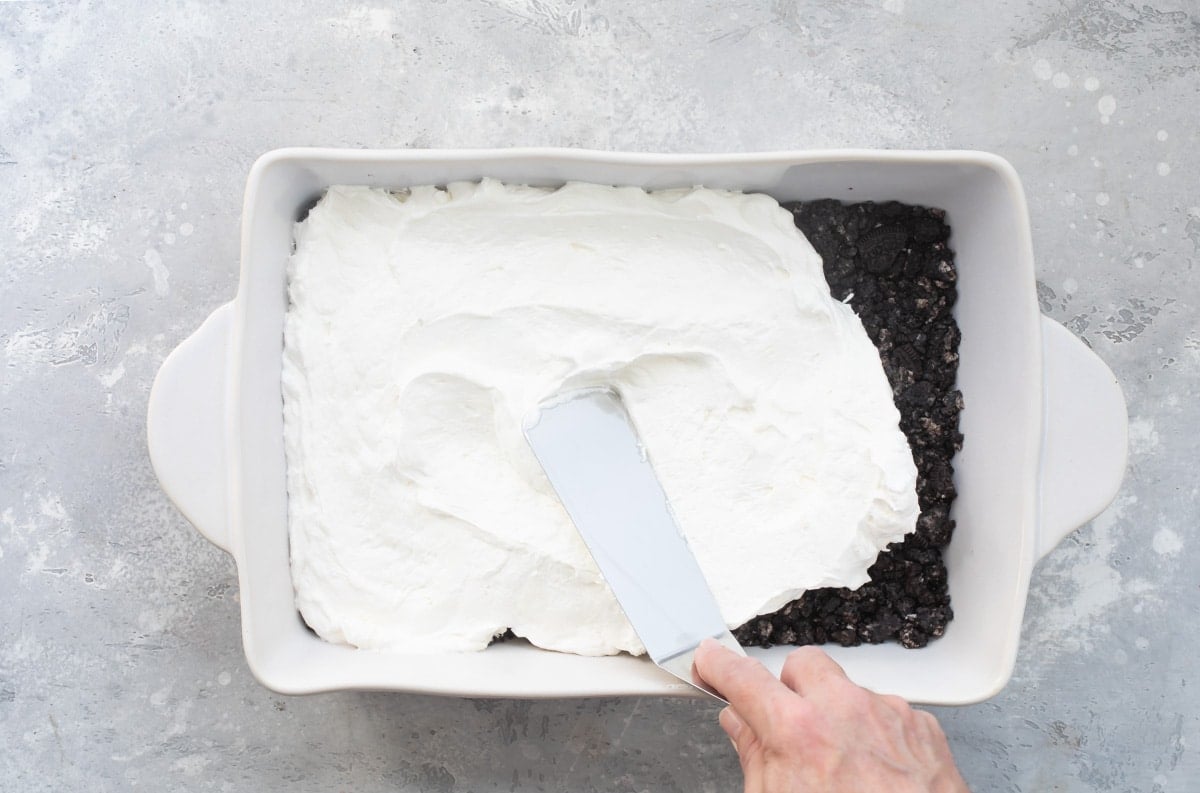 Whipped cream layer of chocolate lasagna being added to a white baking dish.