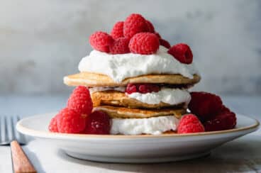 A stack of oatmeal pancakes topped with whipped cream and raspberries on a plate.