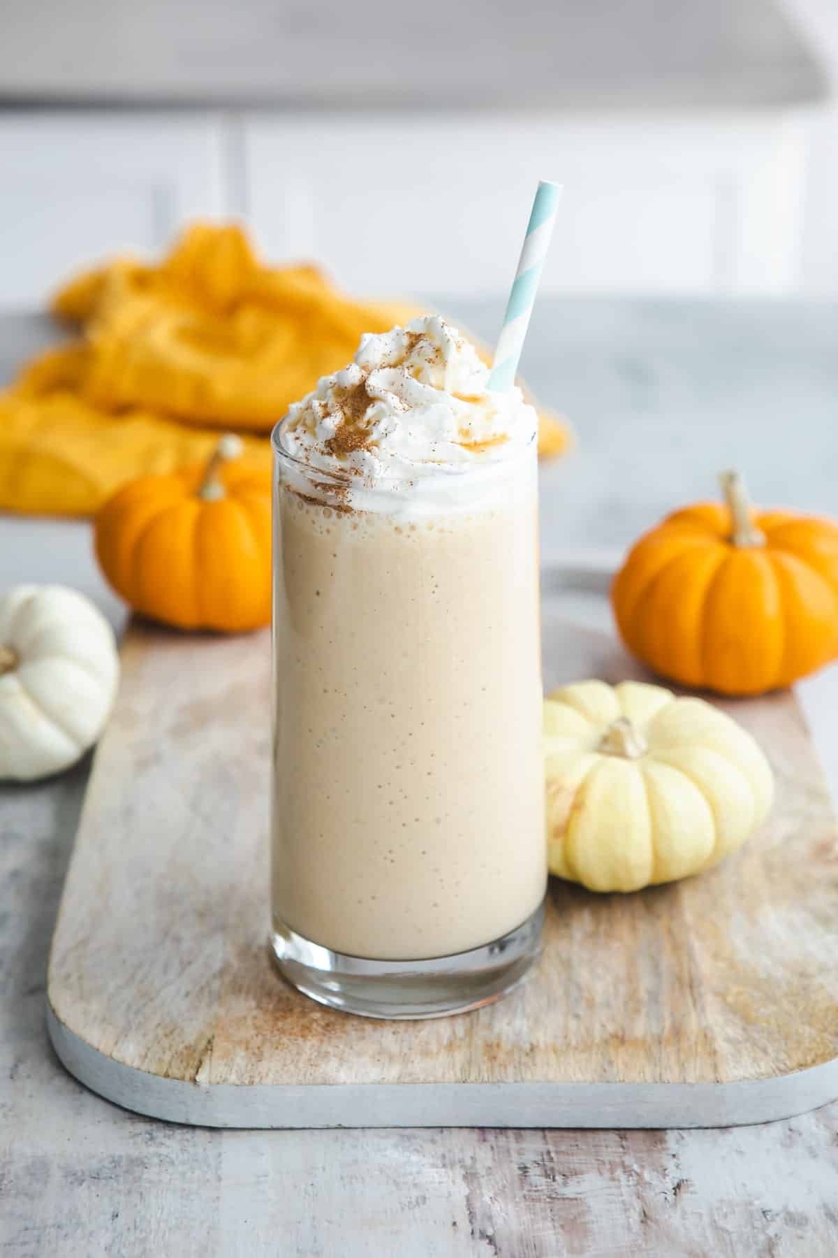 A pumpkin smoothie in a clear glass surrounded by small pumpkins.