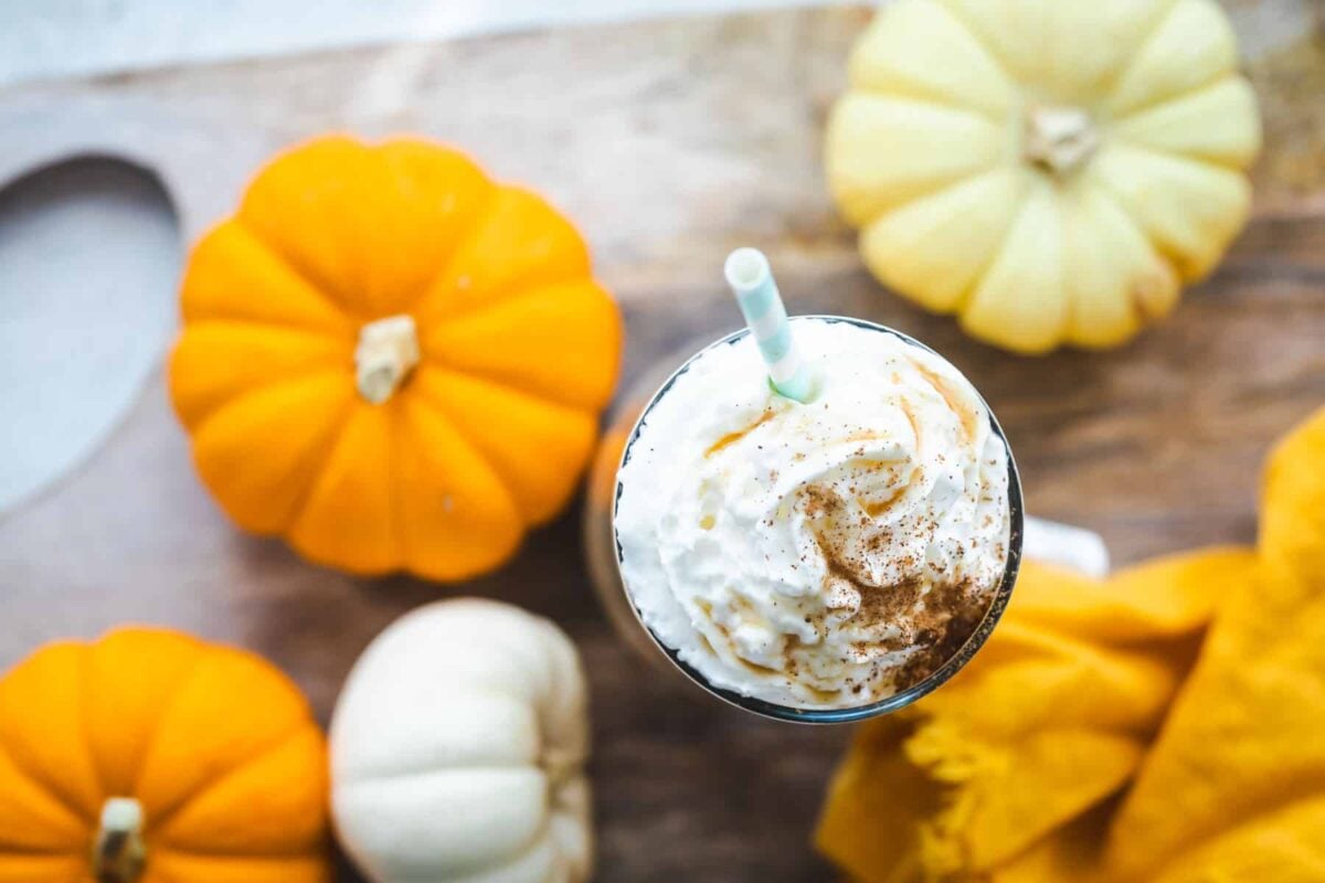A pumpkin smoothie in a clear glass surrounded by small pumpkins.