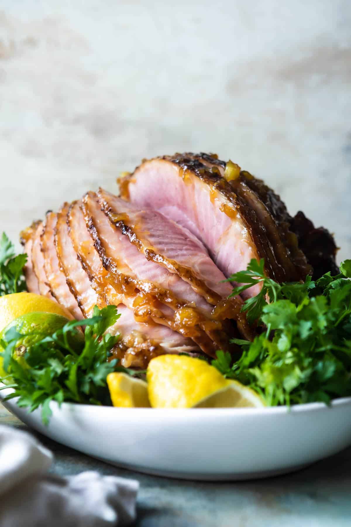 A glazed ham on a platter with fruit and herbs.