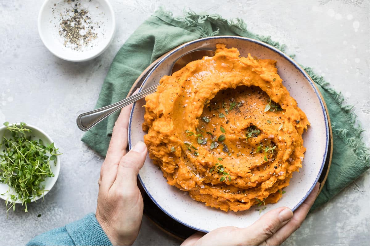 Hands holding a bowl of mashed sweet potatoes with fresh thyme sprinkled on top.