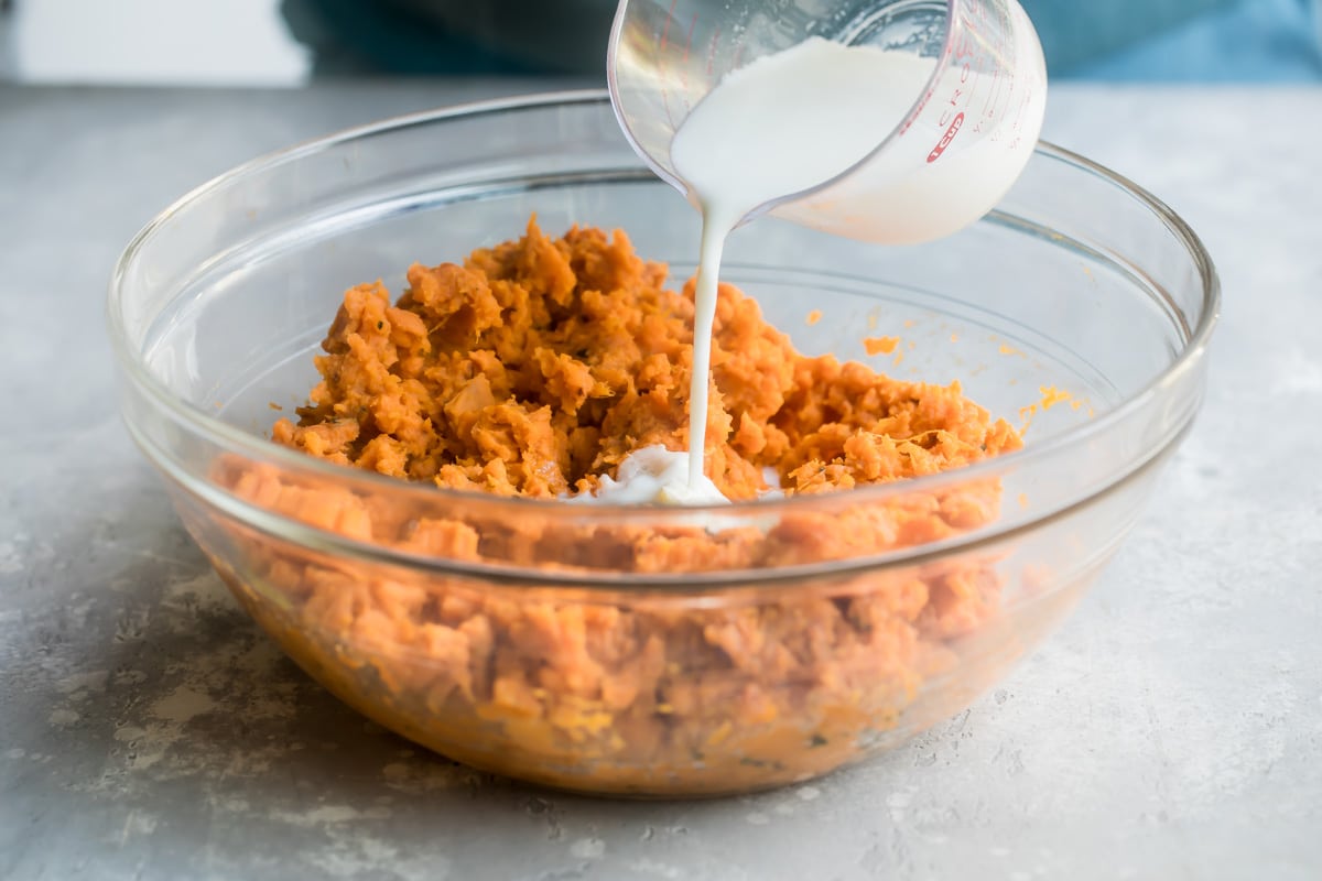 Cream being poured onto mashed sweet potatoes in a clear bowl.