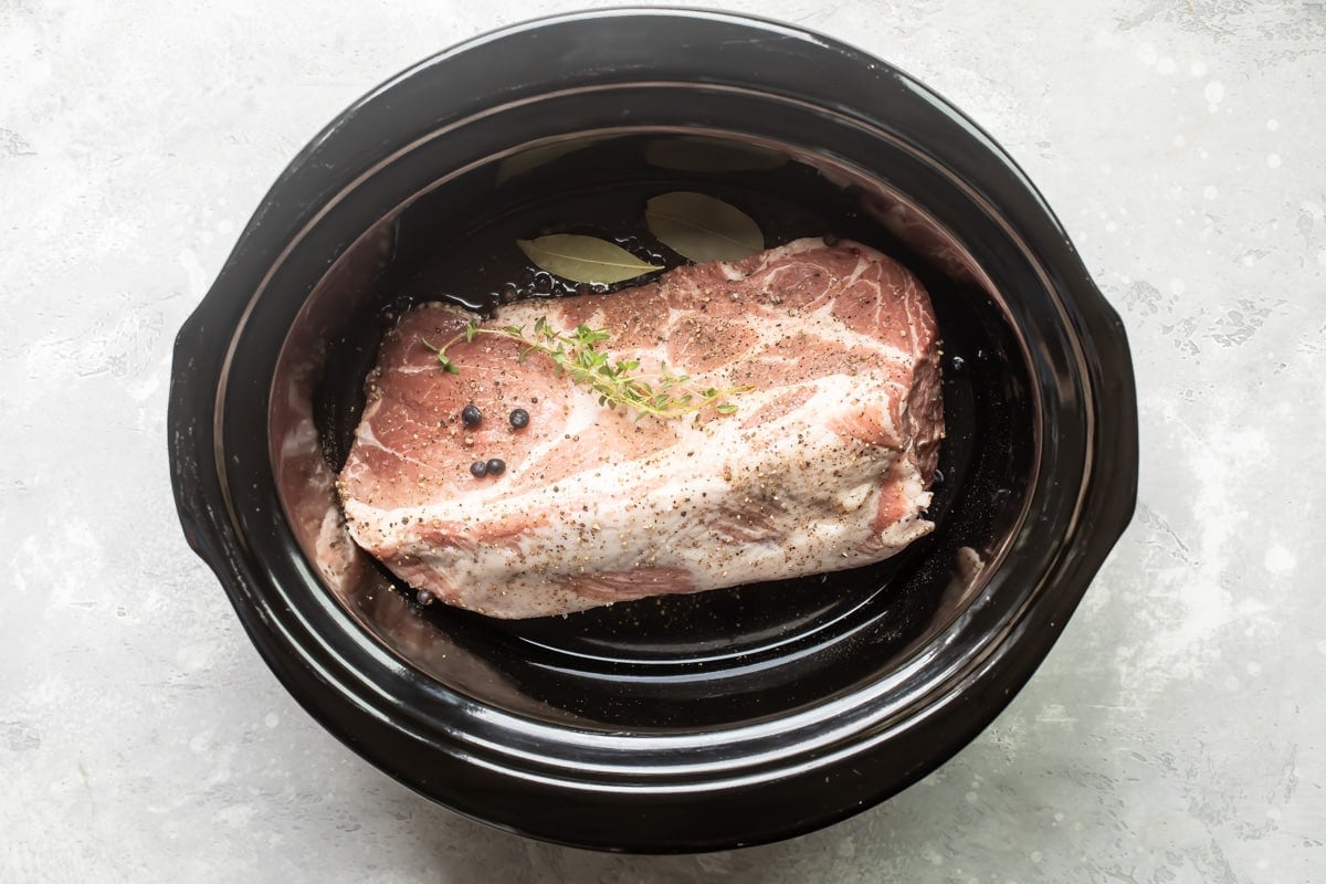 Pork shoulder in the bottom of a slow cooker with juniper berries and a bay leaf.