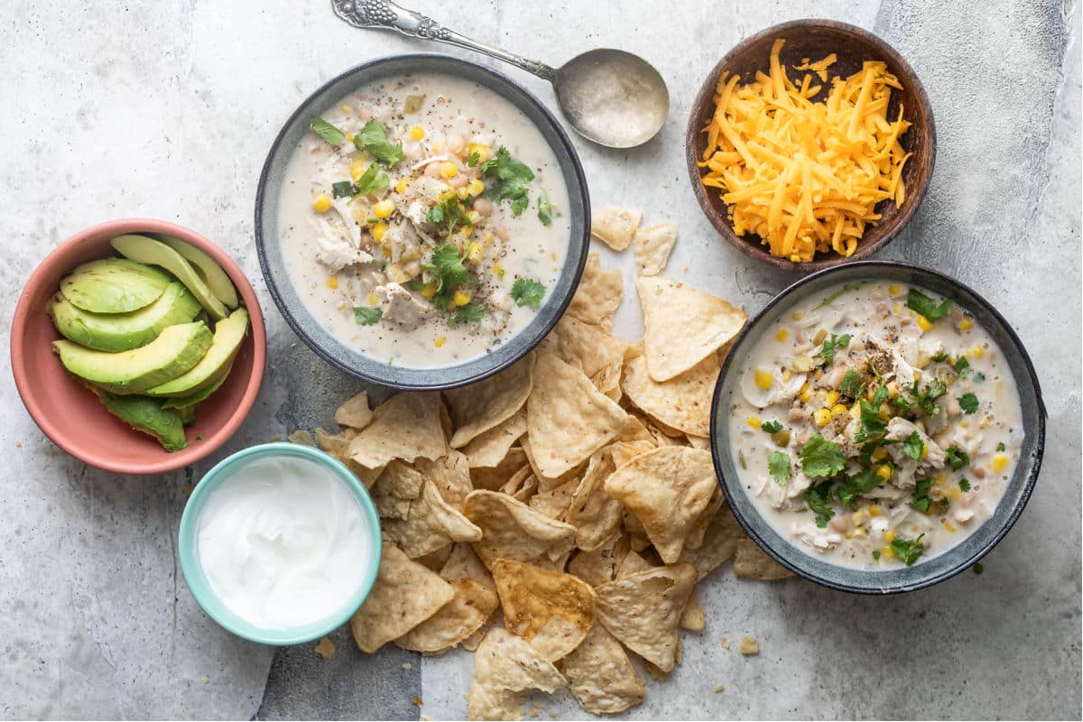 Slow cooker white chicken chili, shredded cheese, avocado slices, sour cream and tortilla chips in various bowls.
