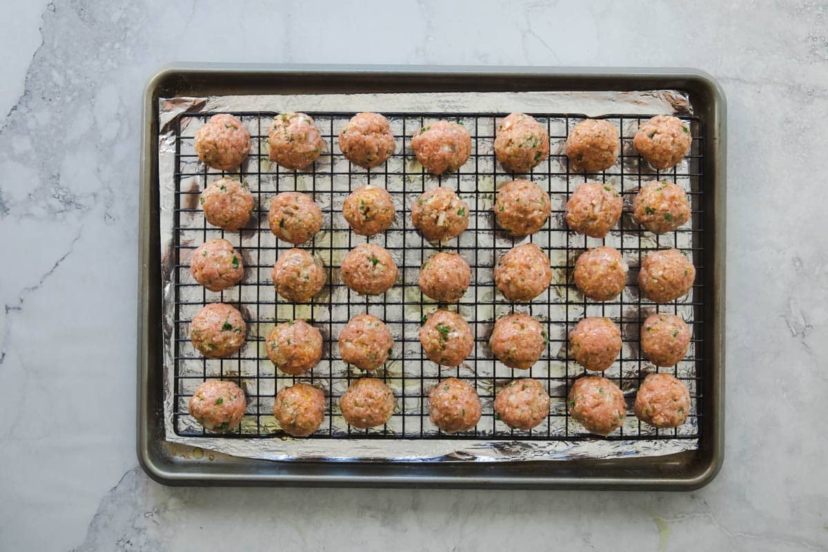 Rows of unbaked meatballs on a baking rack set over a baking sheet.