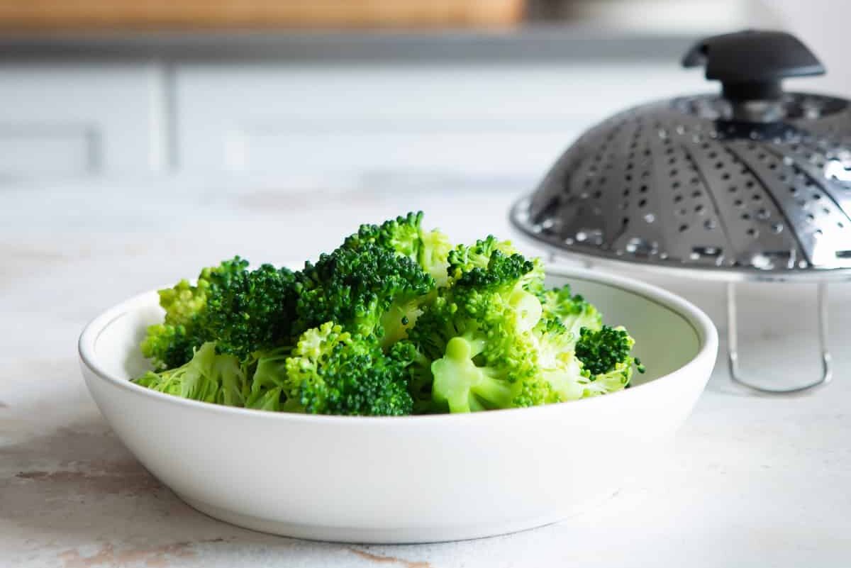A bowl of steamed broccoli next to a steamer basket.