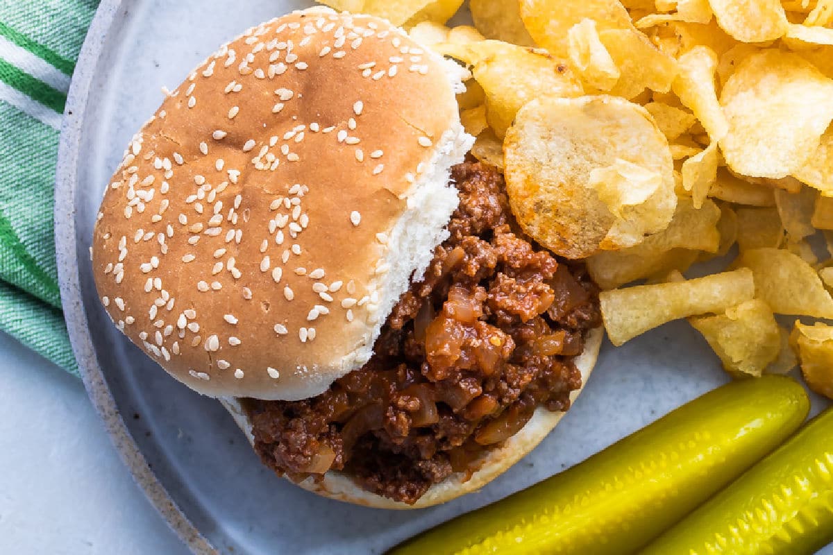 A sloppy joe on a bun with chips and pickles on a blue plate.
