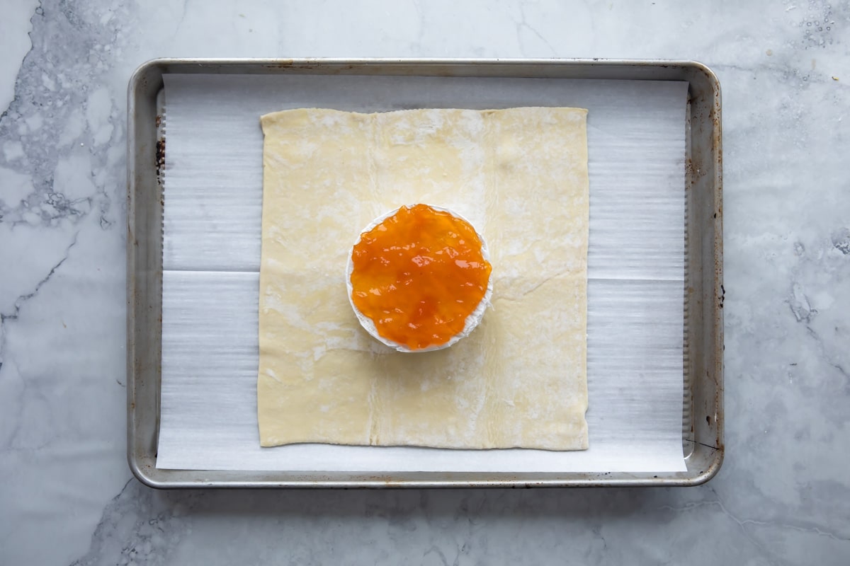 A piece of puff pastry on a baking sheet with a wheel of brie covered in apricot jam on top.