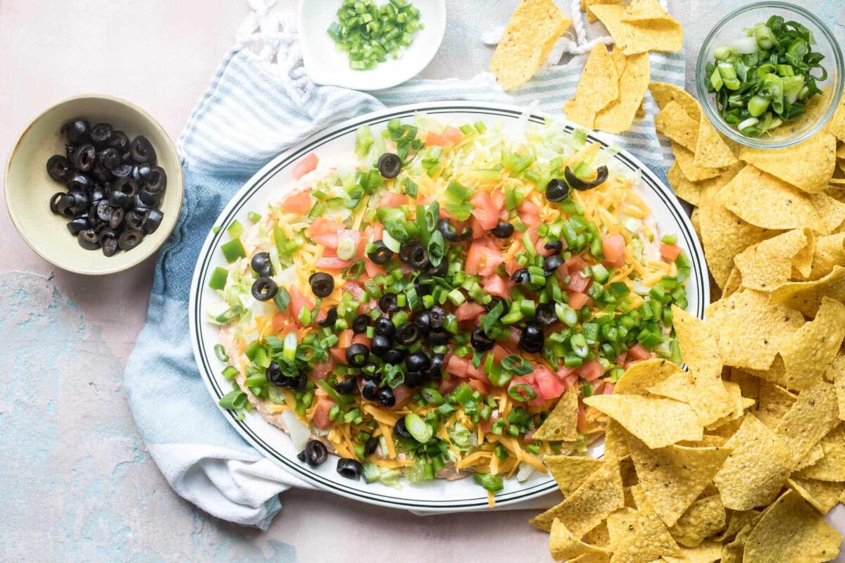 A platter of taco dip surrounded by tortilla chips and small bowls of toppings.