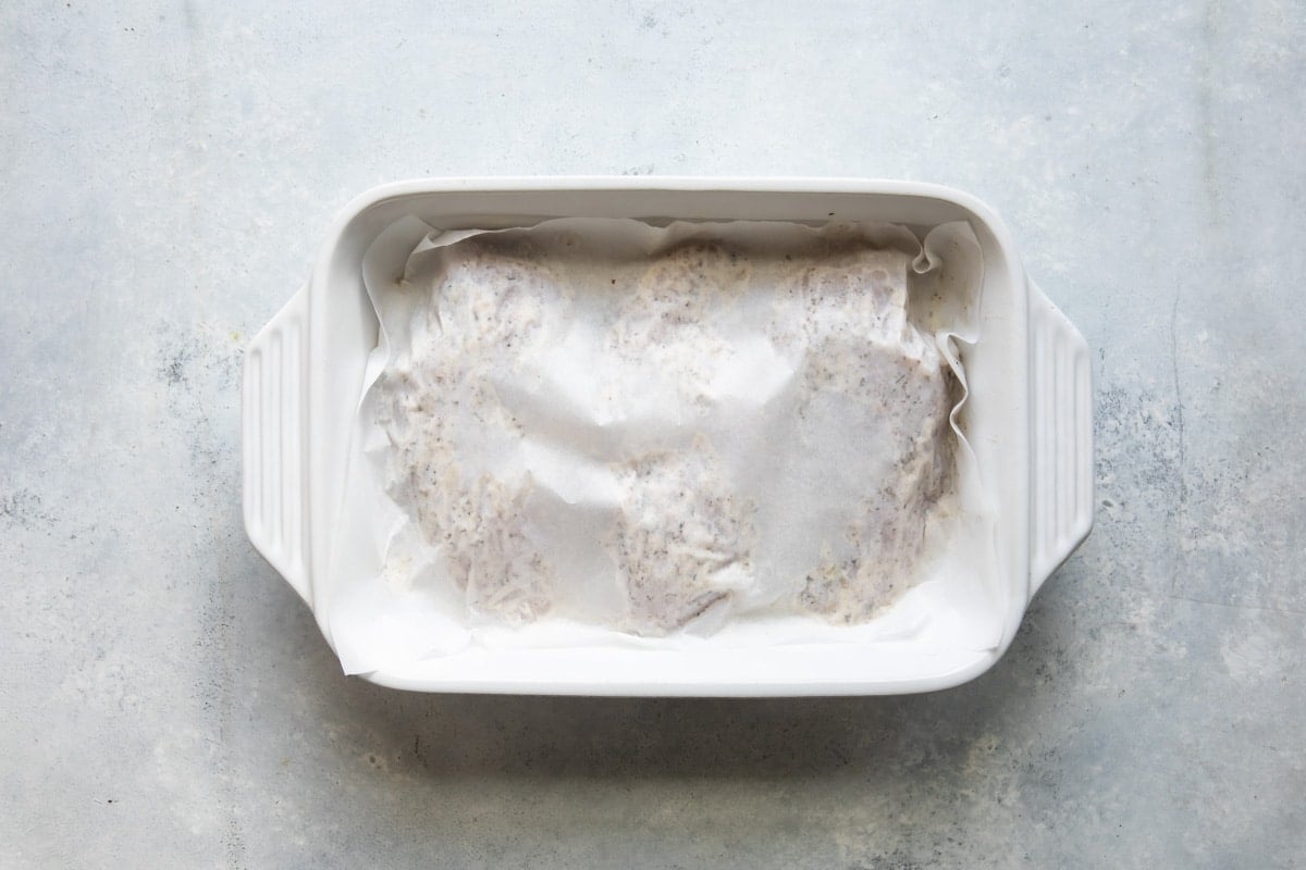 Raw Greek chicken in a baking dish covered in parchment.