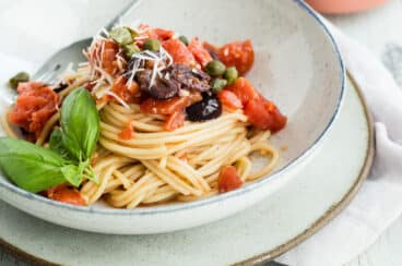 A bowl of Spaghetti alla Puttanesca on a table next to garlic bread.